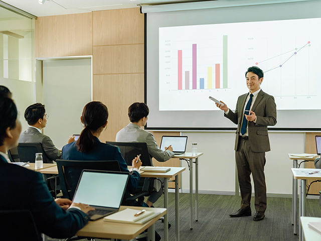 スクリーンにグラフを映し、受講者の前で説明するスーツ姿の講師