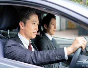 車を運転しているスーツ・ネクタイ姿の男性と、助手席に座るスーツ・ネクタイ姿の男性