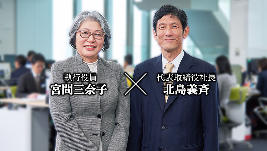 職場フロアーで撮影。左に宮間役員・右に北島社長が並んで笑顔で立っている。