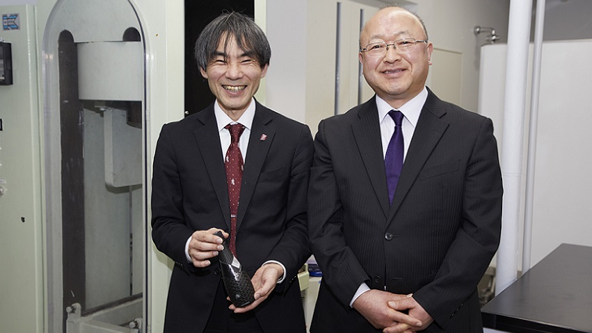 実験用の圧力機の前に立つ、伊藤教授（左）とDNP包装事業部の高橋さん(右)