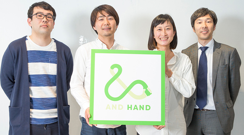 AND HANDの大きなマークを掲げたメンバー4人の集合写真